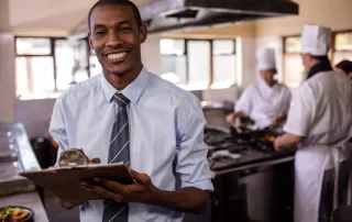 person checking list in front of three chefs in a kitchen